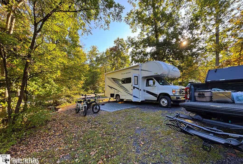 Motorhome and bicycles at a campsite surrounded by trees in early autumn, with a truck bed full of camping gear.