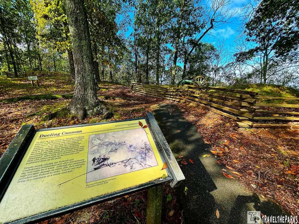 Outdoor historical site with trees, an informational sign titled "Dueling Cannons," and a cannon behind a wooden fence.