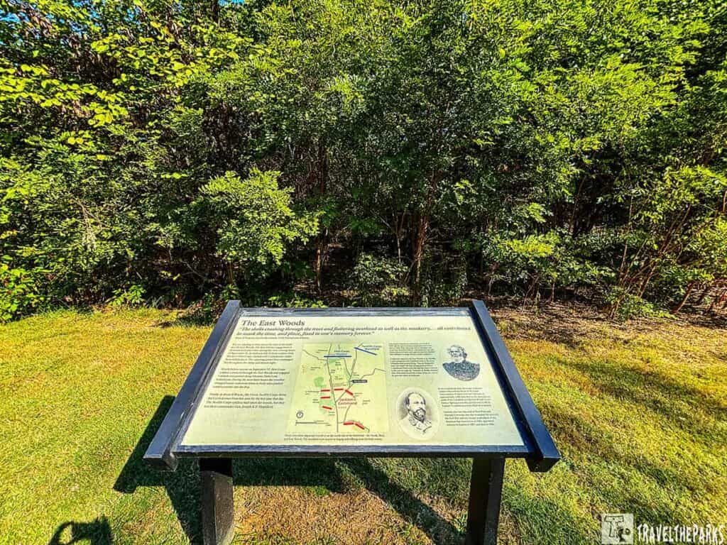Informational sign about "The East Woods" with text, a map, and illustrations, surrounded by grass and dense foliage.