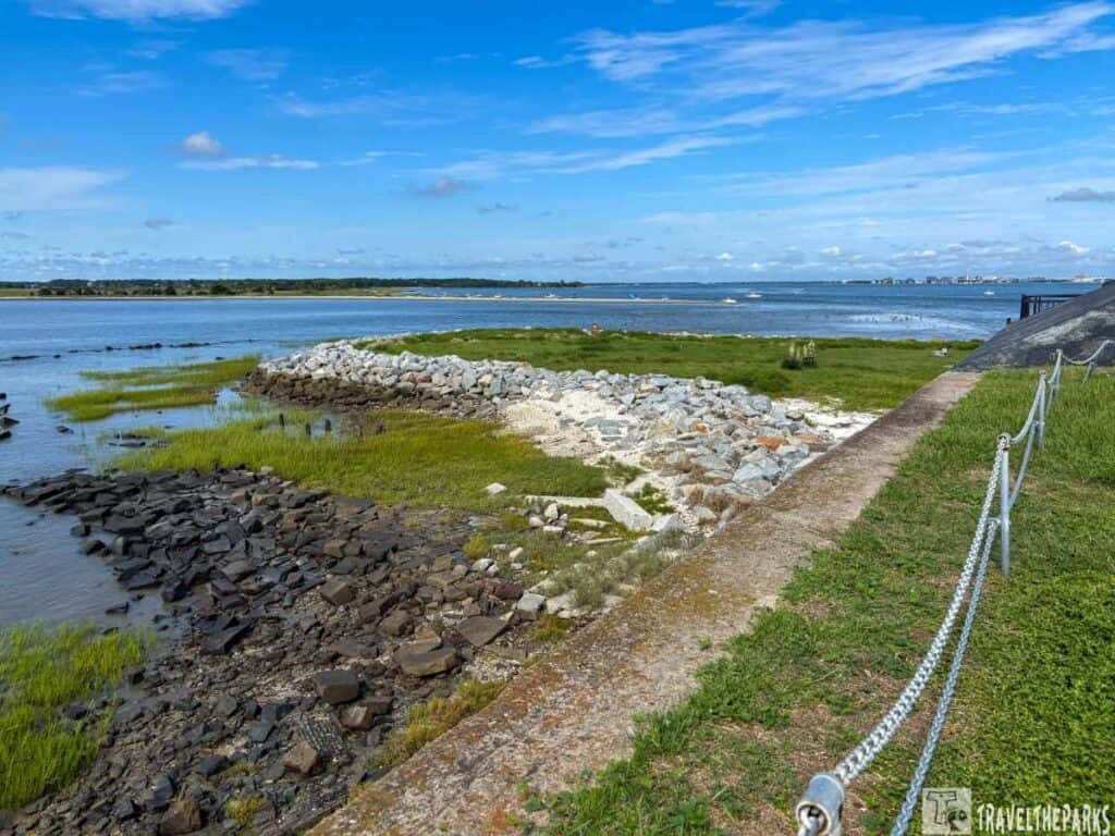 Coastal scene at Fort Sumter with grassy areas, rocky shoreline, and water under a blue sky.