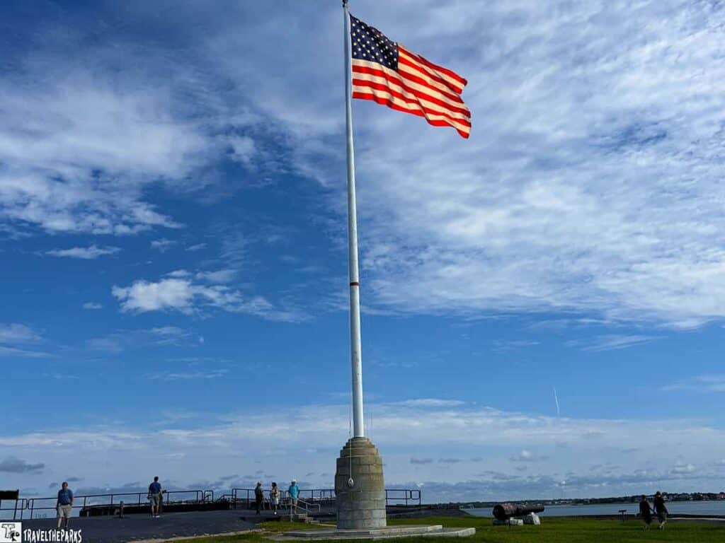 American flag on a flagpole at Fort Sumter with people and ocean in the background.