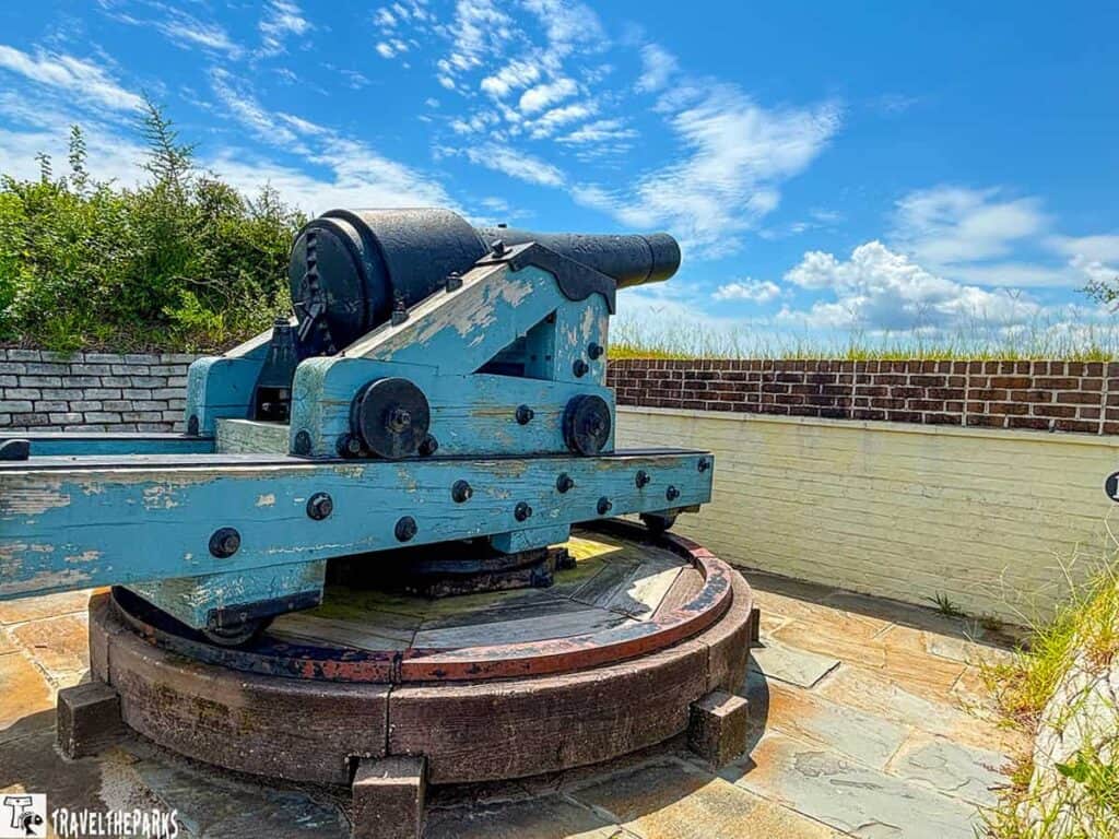 Old blue cannon on a swiveling platform with a brick wall and blue sky in the background.