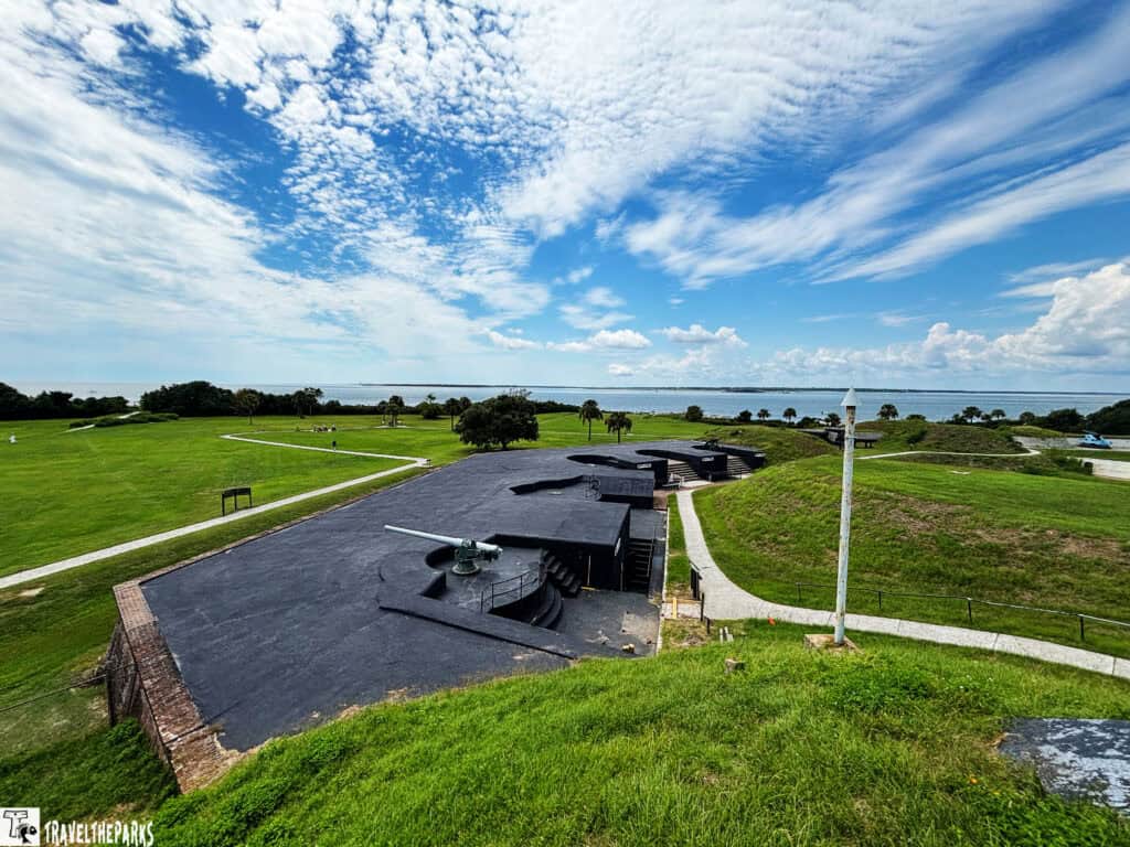 View of Fort Moultrie with a black rooftop and cannon, surrounded by green grass, pathways, trees, and a background of water under a cloud-filled sky.