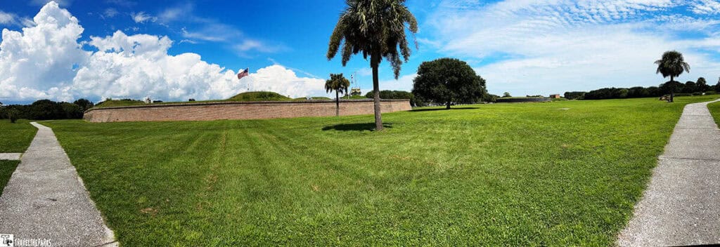 A scenic view of a grassy field with palm trees, brick fort walls, and bright blue sky.

