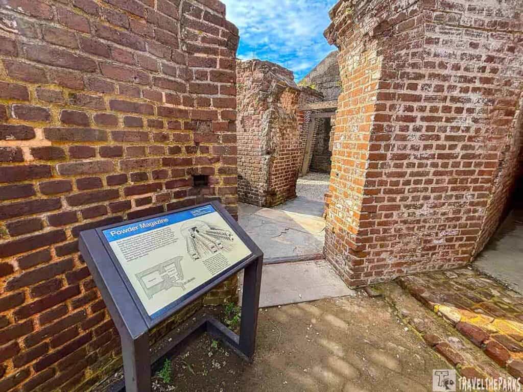  In the foreground, there is an angled informational display sign labeled “Powder Magazine.” The sign includes text and diagrams related to the structure. Surrounding the sign are weathered brick walls, with distinct reddish-brown and yellow hues. The bricks are laid in a consistent pattern, giving a textured appearance. Behind the sign, a narrow passageway between the brick walls leads to a gravel-floored courtyard. Above, the sky is partially visible, showing a bright blue tone with scattered clouds. The ground is a mixture of dirt and stone pavement.