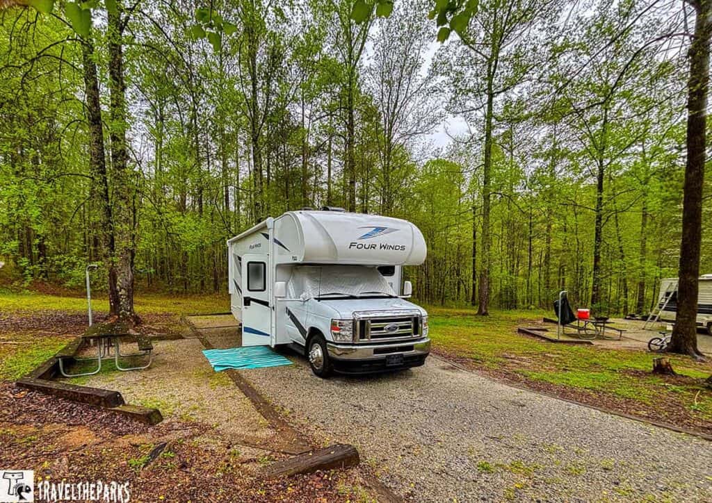 A white RV parked at a forested campsite with a picnic table and another RV in the background.