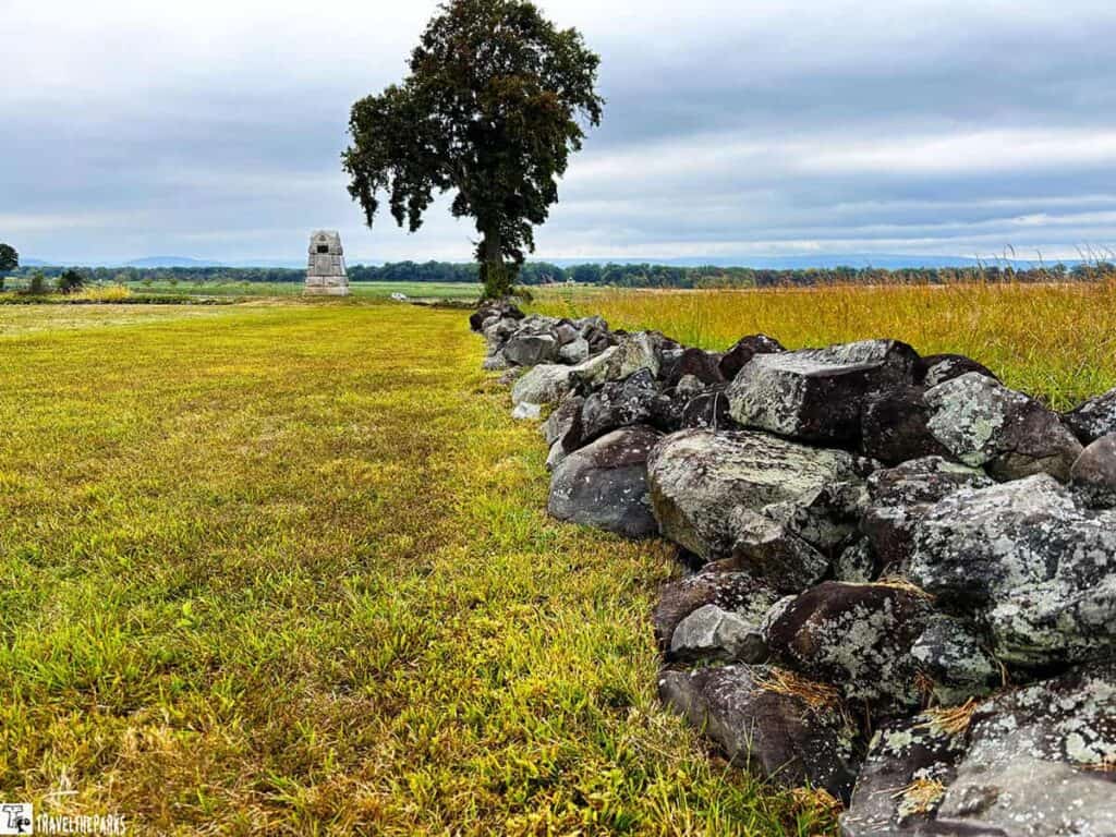 Stone wall and field at Gettysburg National Military Park, with a tree and monument in the background under a cloudy sky.