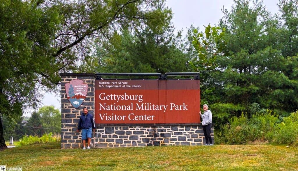 Gettysburg National Military Park Visitor Center sign with stone and wood, flanked by two people, set against a backdrop of trees.