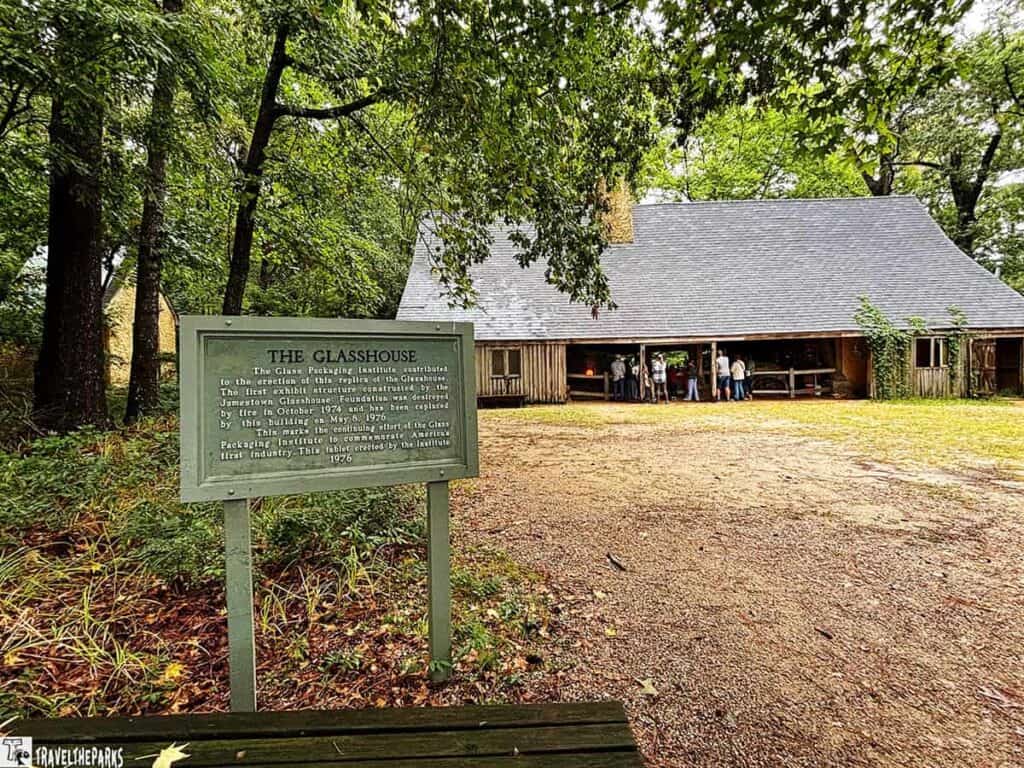 A historic site at Jamestowne featuring a replica of the Glasshouse with an informational sign in the foreground. People are gathered under the building's shelter.