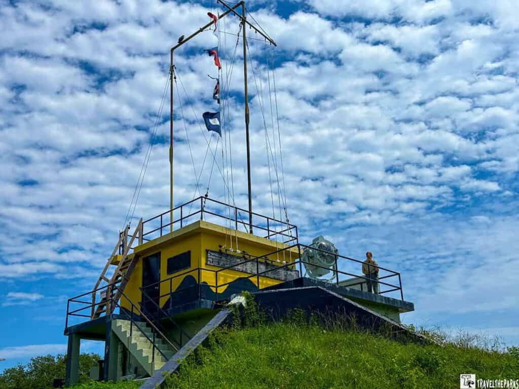 harbor entrance control post at Fort Moultrie. The building is rectangular and painted in yellow with a dark blue wave pattern near the bottom. It is surrounded by metal railings. Several flagpoles rise from the roof, with various maritime flags fluttering against a partly cloudy sky. A staircase on the left leads up to the building, and a spotlight is mounted on the roof next to a person standing near the railing. The sky is bright blue with scattered cumulus clouds, adding a picturesque backdrop.