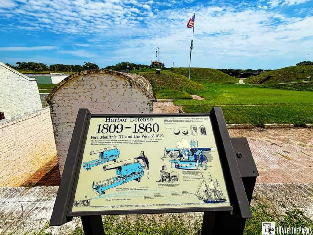 Informational sign at Fort Moultrie about harbor defense from 1809 to 1860 with grass mounds and an American flag in the background.