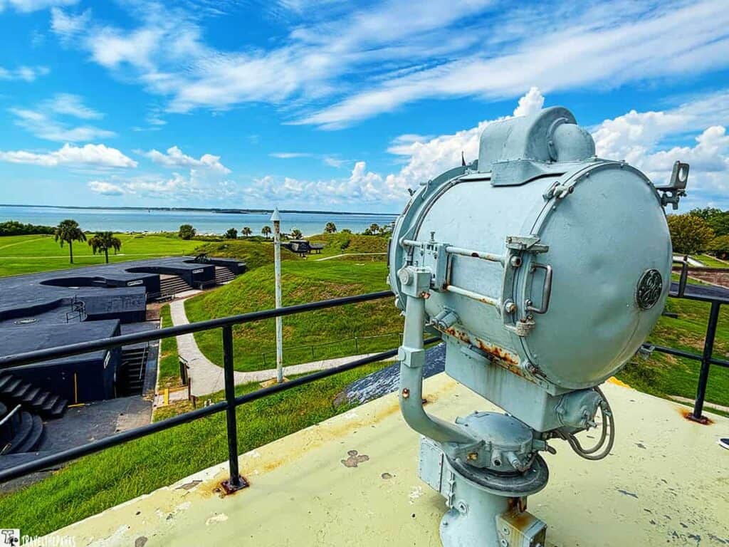A large metal spotlight on a rooftop at Fort Moultrie with grassy areas, palm trees, and the ocean in the background.

