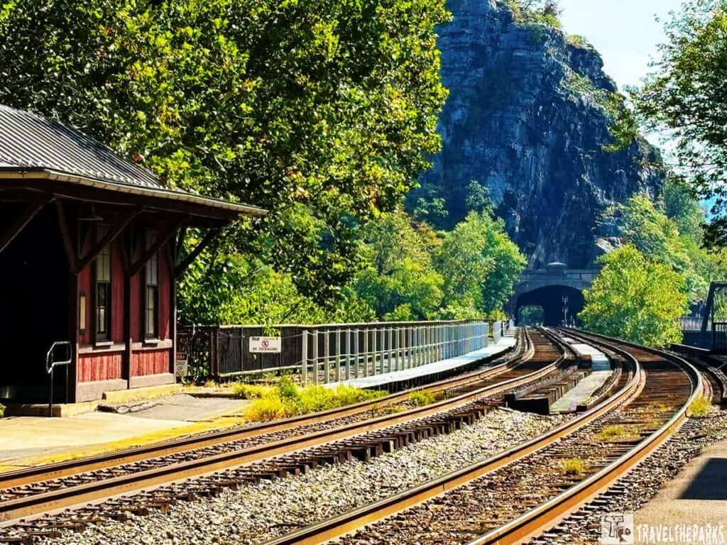 Curved railway tracks leading into a tunnel surrounded by trees and a rock formation, with a red train station building on the left.