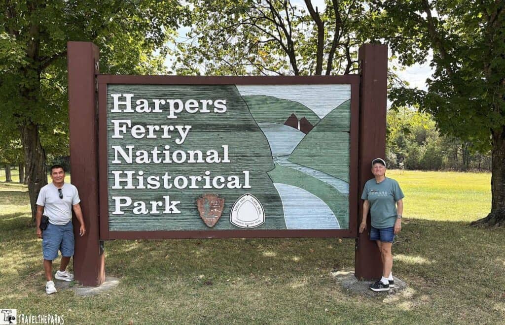 Two people standing by the Harpers Ferry National Historical Park sign with trees in the background.