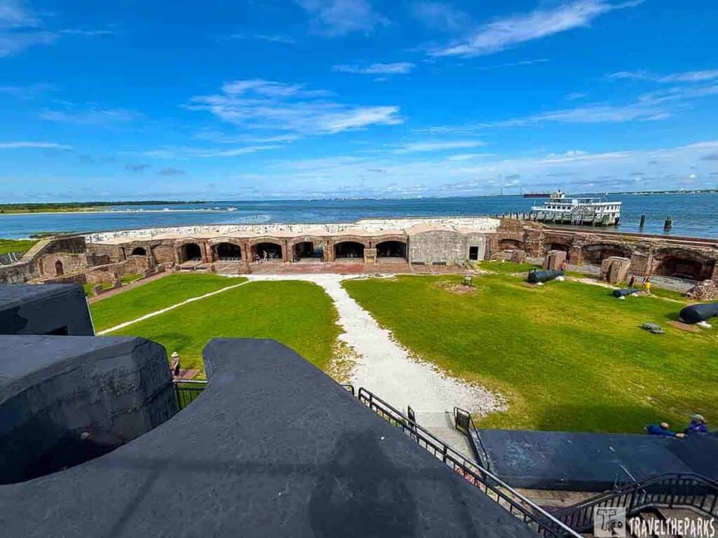 Aerial view of Fort Sumter showing a grassy courtyard, brick structures, cannons, and a ferry on the water.
