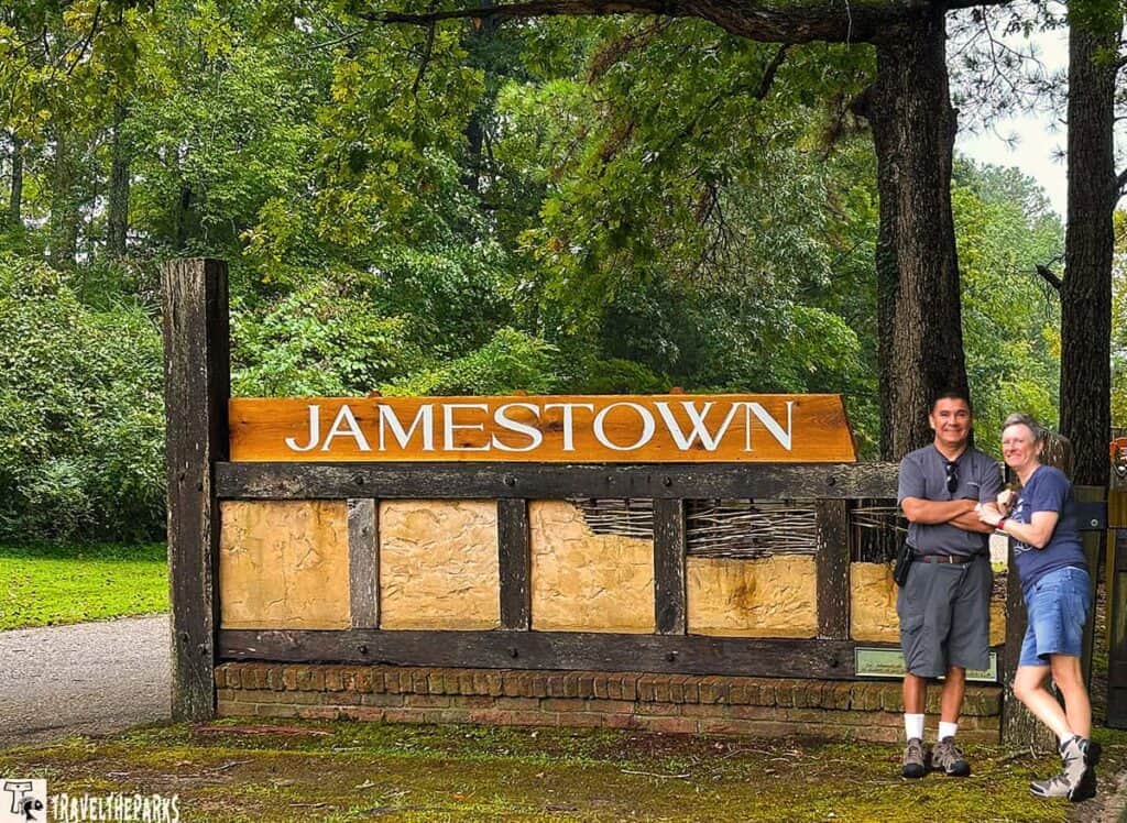 A wooden sign reading "JAMESTOWN" in white letters with greenery in the background and two people standing beside it.