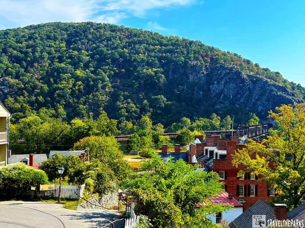Scenic view of Harper's Ferry with historic buildings, a mountain, and a railway bridge beneath a clear blue sky.