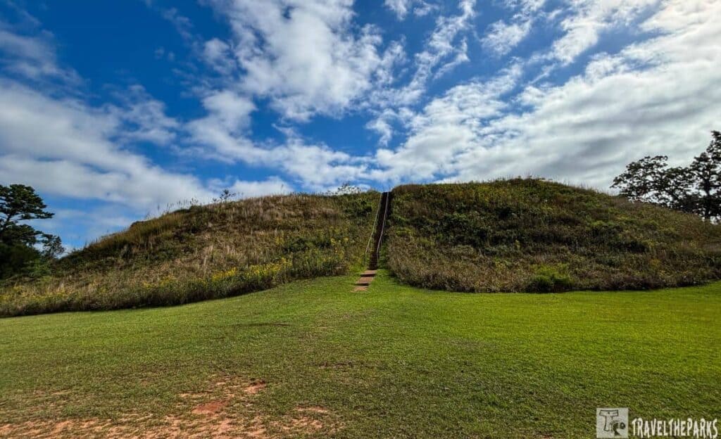 A grassy mound A Kolomaki Mounds State Park with a wooden staircase under a blue sky with clouds.

