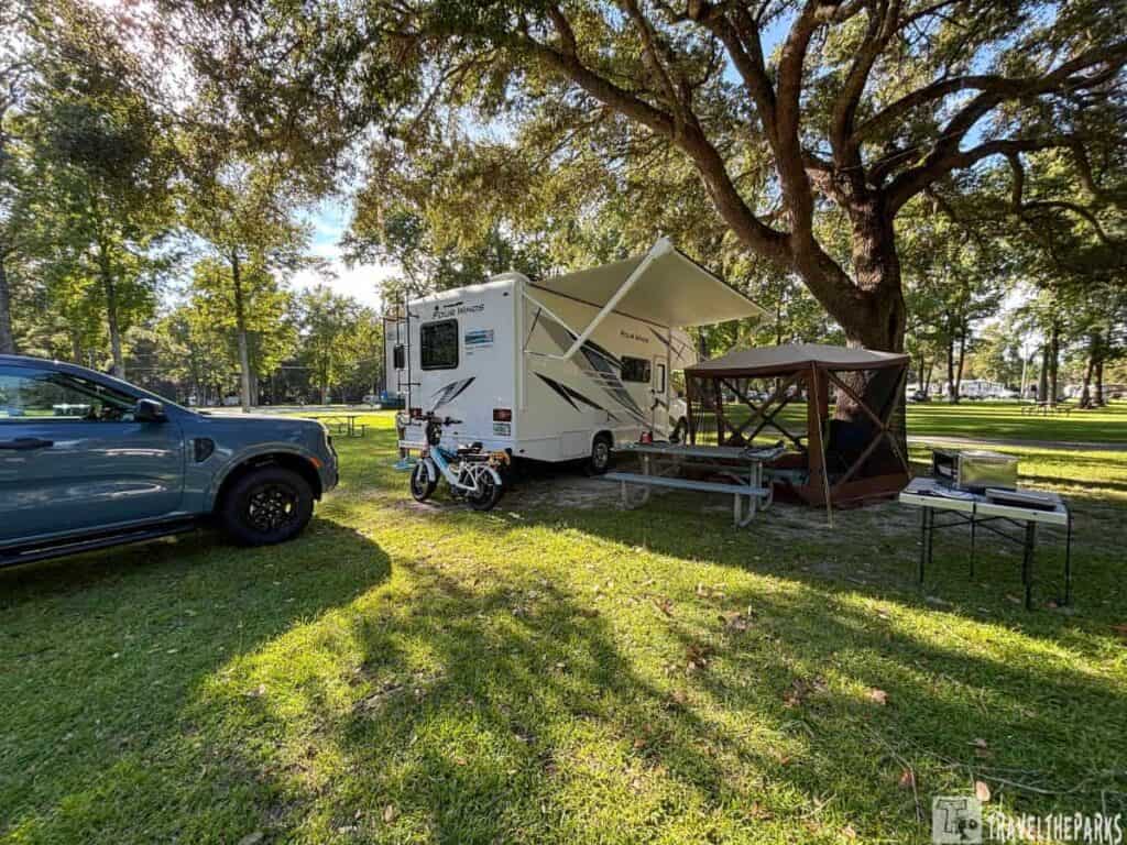 A campground scene with an RV, a small gazebo, and a blue vehicle under large oak trees.