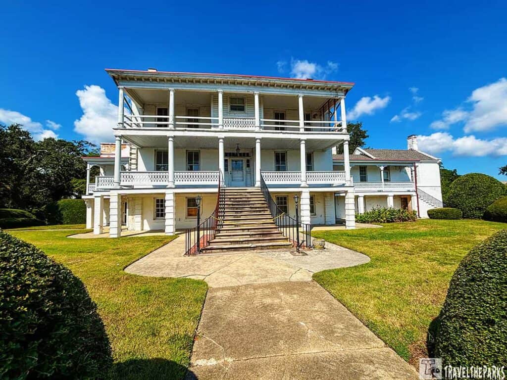 Historic white three-story building with porches and columns, wide staircase, and surrounding greenery under a blue sky.