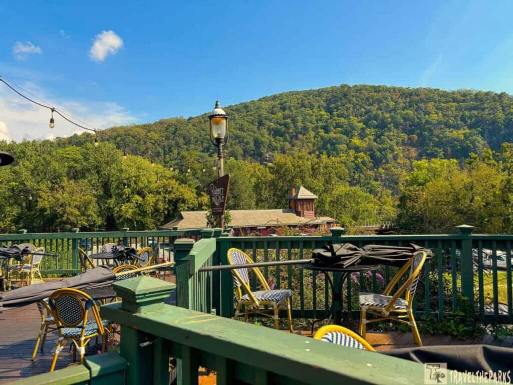 An outdoor patio with tables and chairs overlooks a lush green hillside under a blue sky. A sign reads 