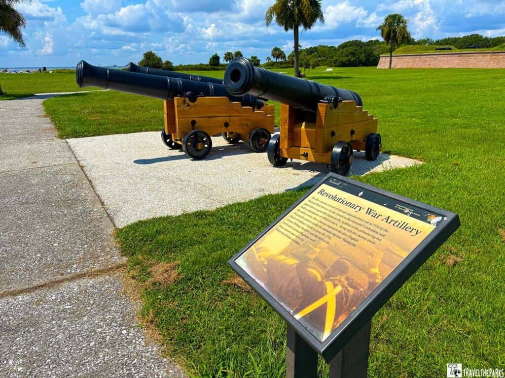 Two historical Revolutionary War cannons on wooden carriages at Fort Moultrie with an informational plaque in the foreground, set against a green landscape with palm trees and a brick wall.

