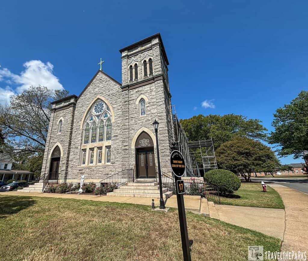 Gothic-style stone church with a rose window, surrounded by a lawn and trees under a clear blue sky.