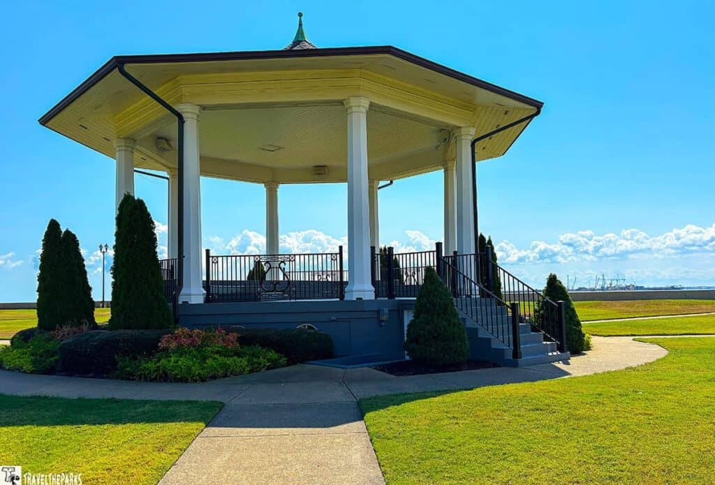 An octagonal bandstand with a wide roof and tall white columns, surrounded by grass and shrubs.