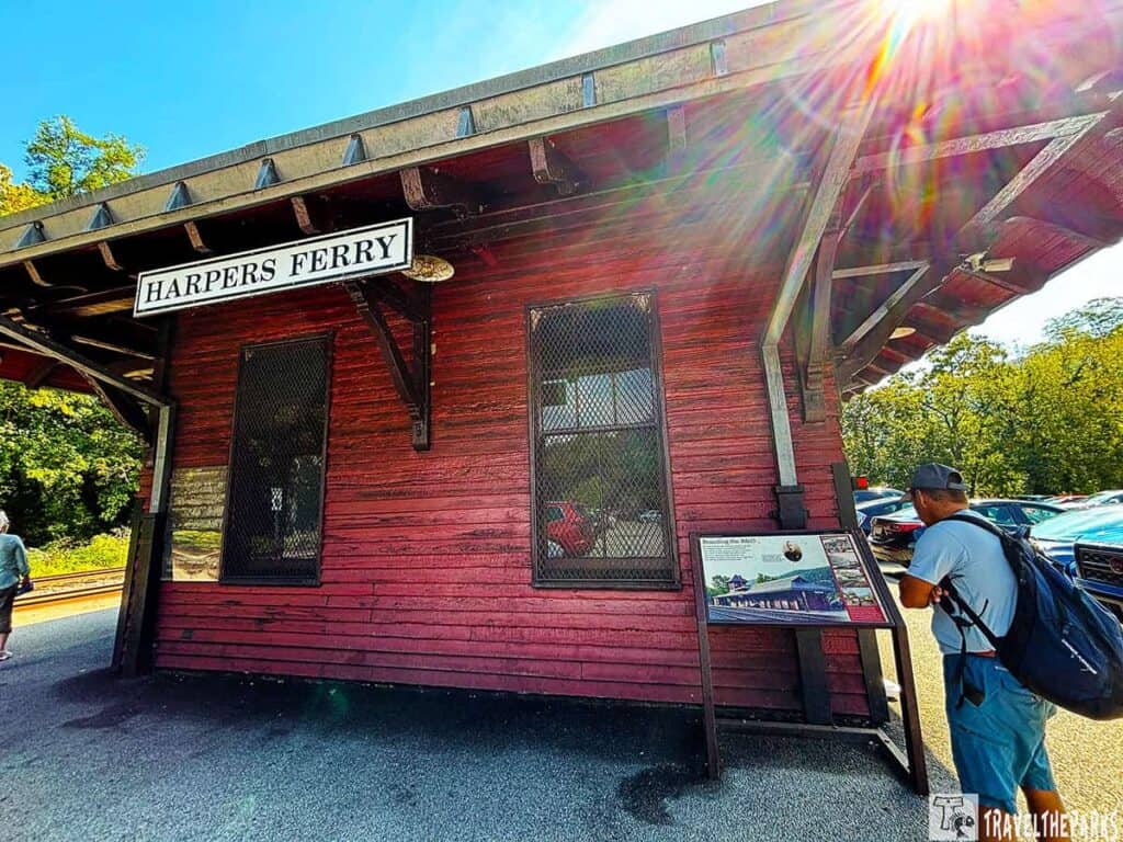 Exterior of Harpers Ferry Train Station with red wooden siding, sign, lens flare, and a person reading an informational display.