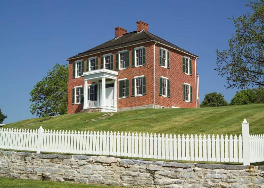A historic Pry house with green shutters on a grassy hill behind a white picket fence and stone wall.