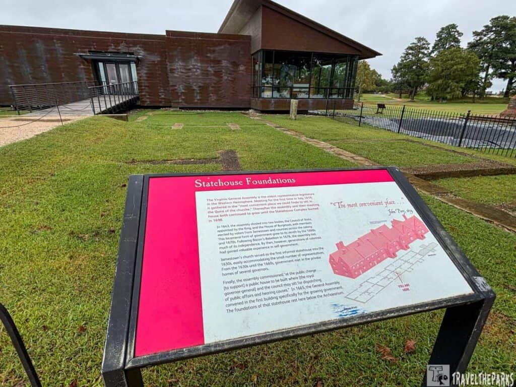 A historical site with a modern museum building and a sign titled "Statehouse Foundations" on a grassy area.