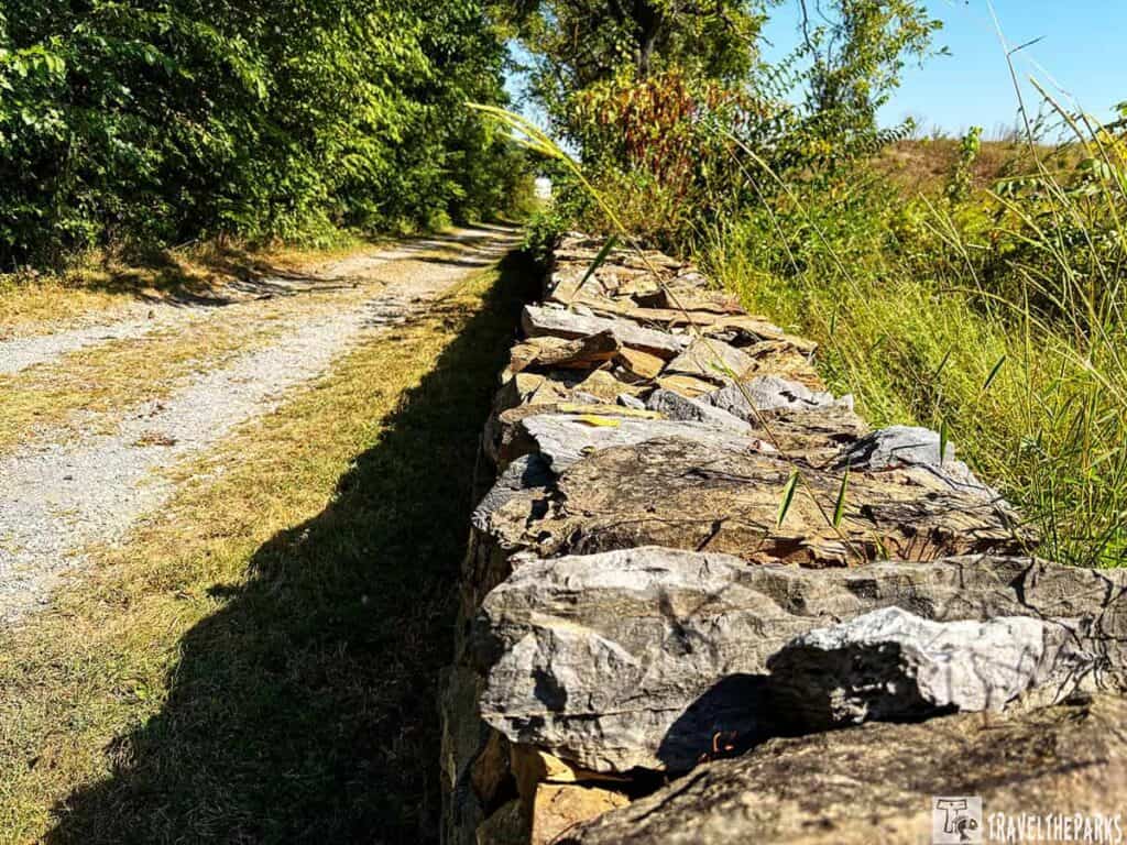 A stone wall next to a gravel path with dense foliage on the left.