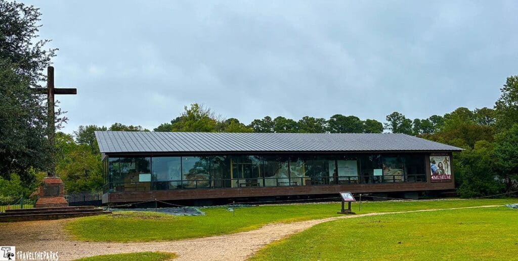 Voorhees Archaearium Museum: a modern glass-fronted building with a metal roof, a wooden cross, and a dirt path, set against an overcast sky.