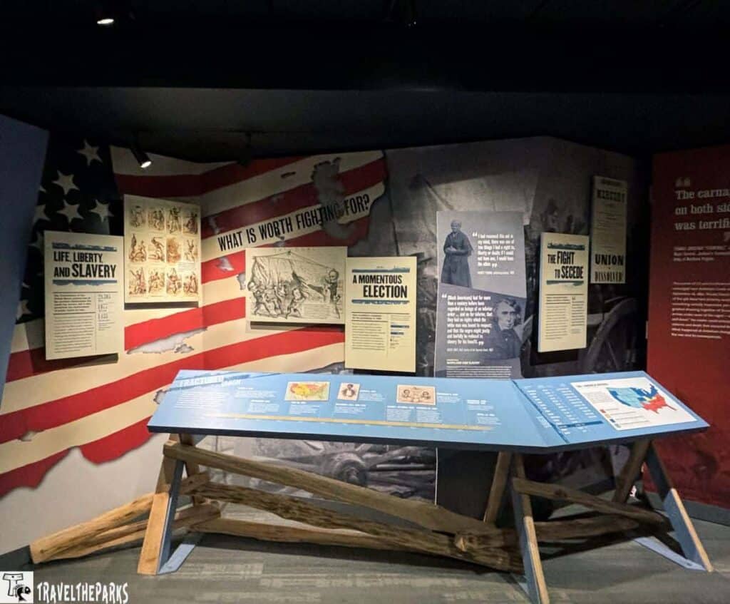 an exhibit at the Antietam National Battlefield visitor center. The backdrop features a large American flag with red and white stripes, slightly tattered in appearance. Attached to the wall are several informational panels with headings like "Life, Liberty and Slavery," "What Is Worth Fighting For?" "A Momentous Election," "The Fight to Secede," and "Union Dissolved." Each panel contains historical texts and images relevant to the Civil War era. A freestanding blue information panel is positioned in front of the wall, supported by wooden stands, featuring additional historical content and a map.