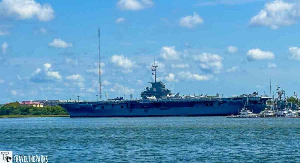 Battleship USS Yorktown docked in a harbor with a cloudy sky and smaller boats nearby.