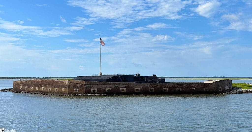 Fort Sumter with a brick structure and an American flag on a tall pole, surrounded by water under a blue sky.