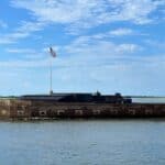 Fort Sumter with a brick structure and an American flag on a tall pole, surrounded by water under a blue sky.