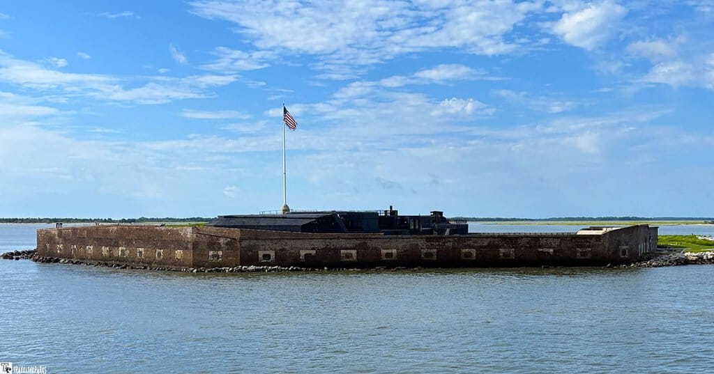 Fort Sumter with a brick structure and an American flag on a tall pole, surrounded by water under a blue sky.