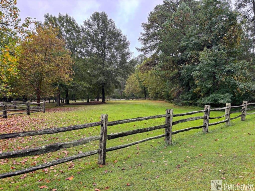 A grassy field Governor Yeardleys 1620s property at Historic Jamestowne with a rustic wooden fence and trees with autumn foliage under an overcast sky.