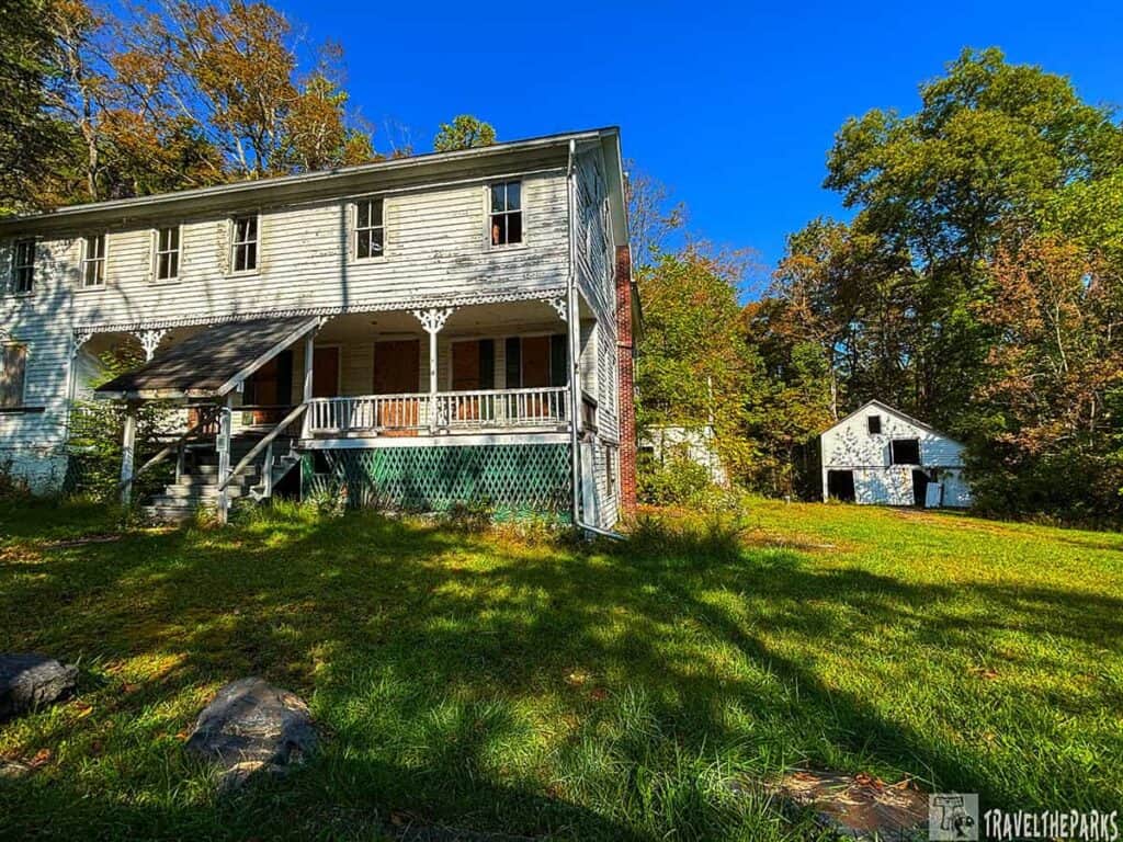 A weathered two-story house with a porch, surrounded by trees, with a blue sky overhead. An outbuilding is nearby.
