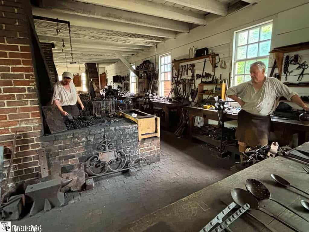 Colonial Williamsburg blacksmith workshop with two individuals in period clothing and various tools.