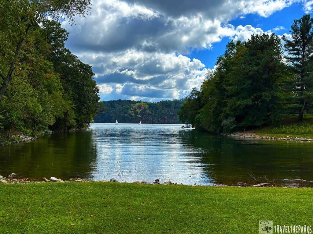 A peaceful lakeside with trees on either side, small sailboats in the distance, and a partly cloudy sky.
