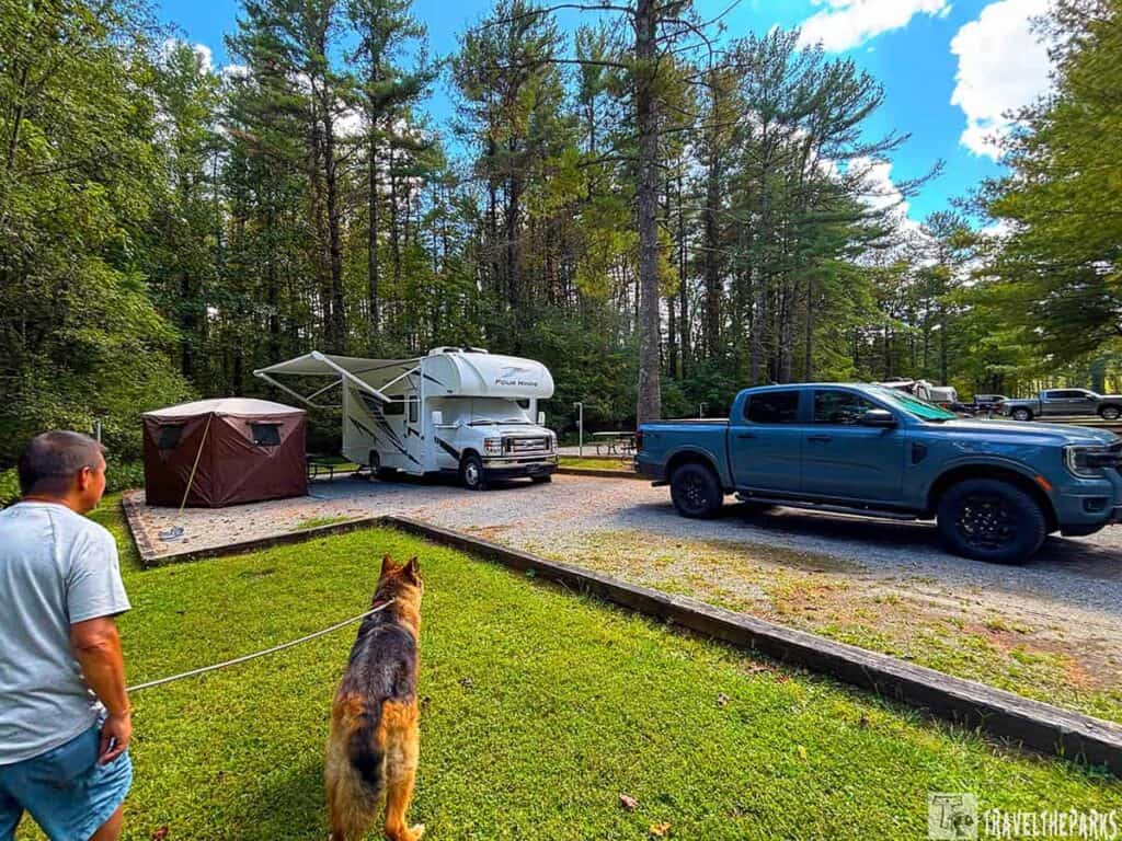 A man and a German shepherd stand on grass near a brown tent, an RV, and a blue pickup truck at a wooded Claytor Lake State Park camping site on a sunny day.