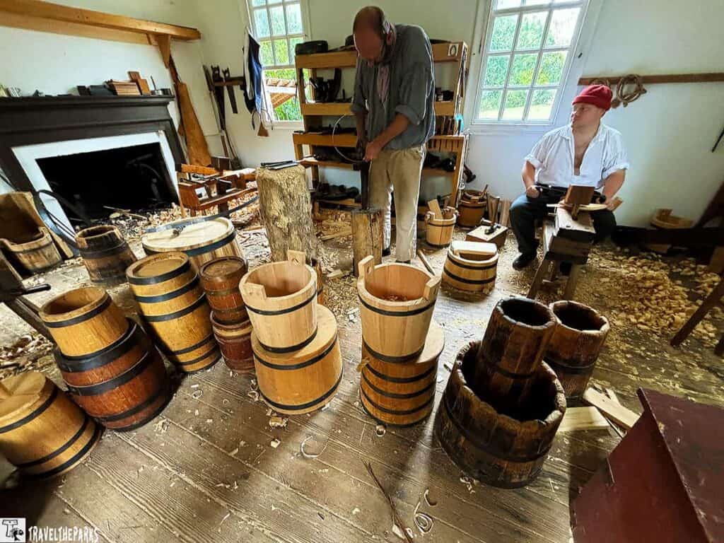 Interior of a cooper's woodworking shop with wooden buckets, barrels, and two craftsmen at work.