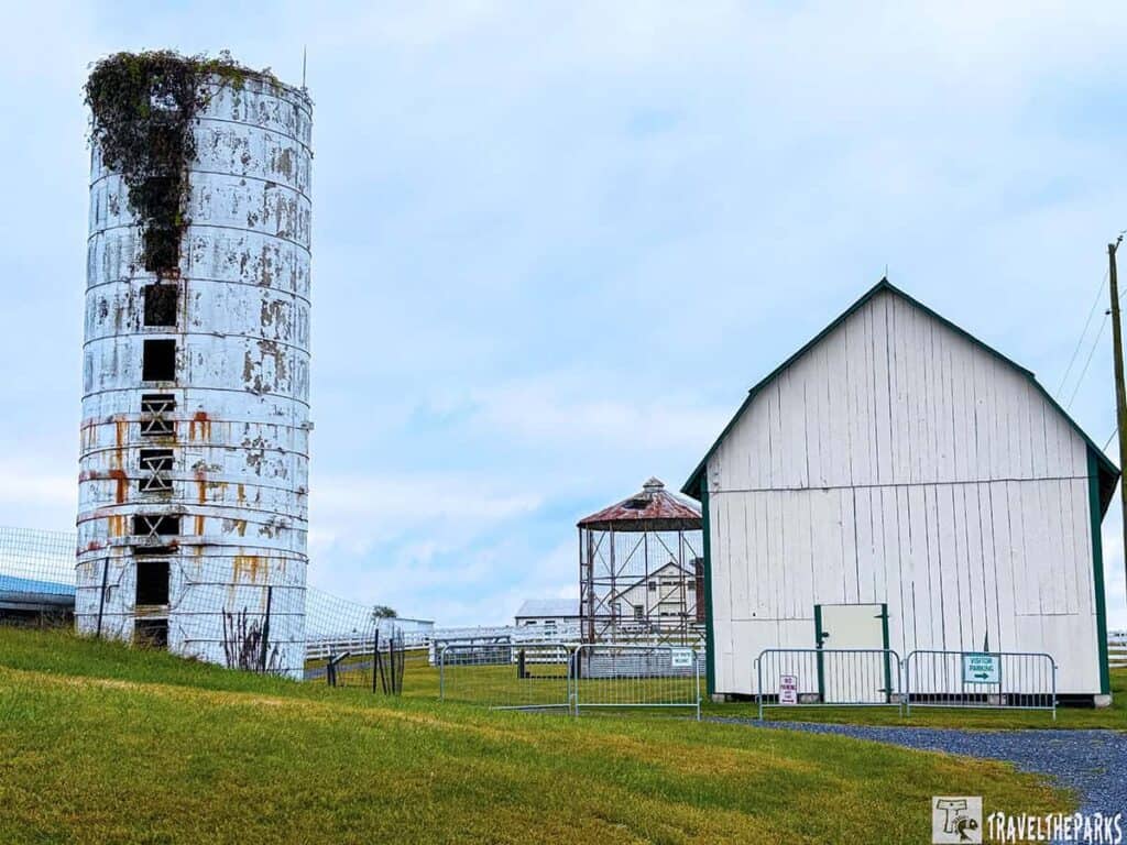 A white cylindrical metal structure with rust and vegetation, next to a large white barn with a green roof on a grassy slope.