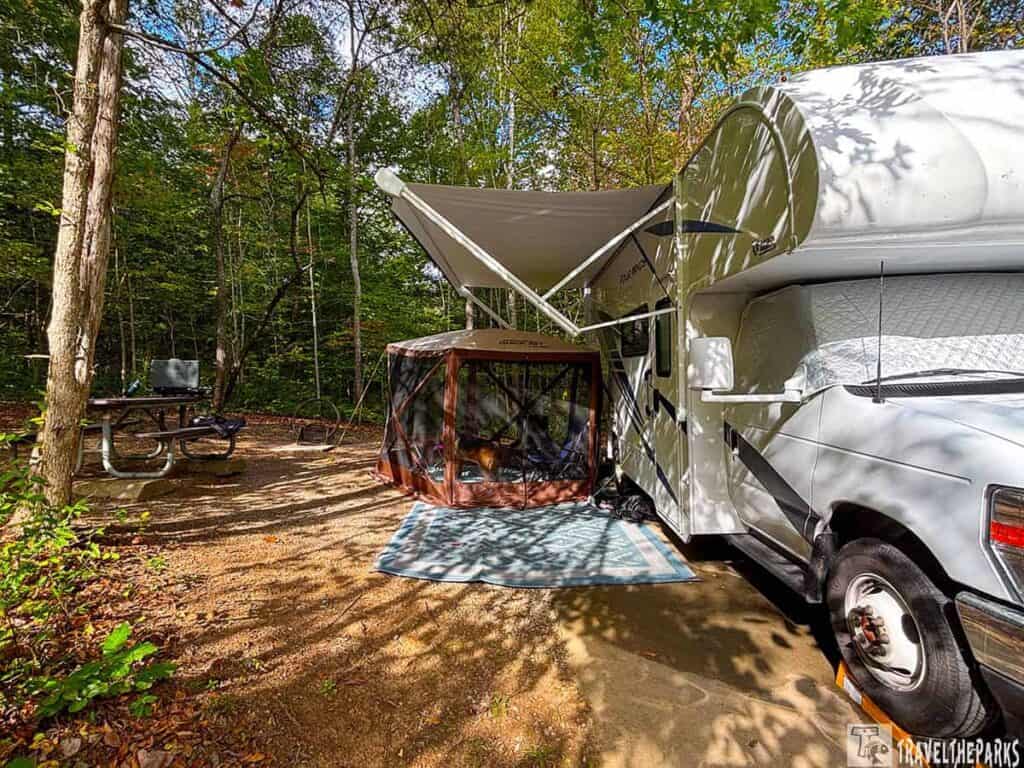 Campsite in a wooded area with a white RV, an awning, a brown screened tent, and a picnic table.