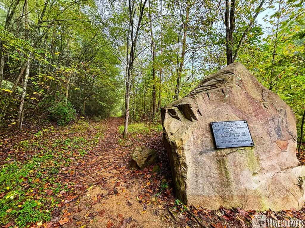 A hiking trail in Cumberland Gap National Historical Park, with a large rock bearing a metal plaque beside the forested path.