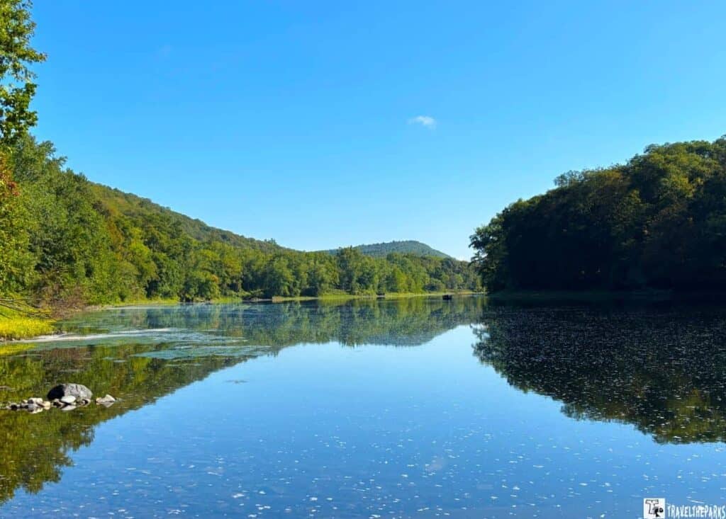 A calm river reflecting trees and a blue sky, surrounded by lush green forests at the Delaware Water Gap.

