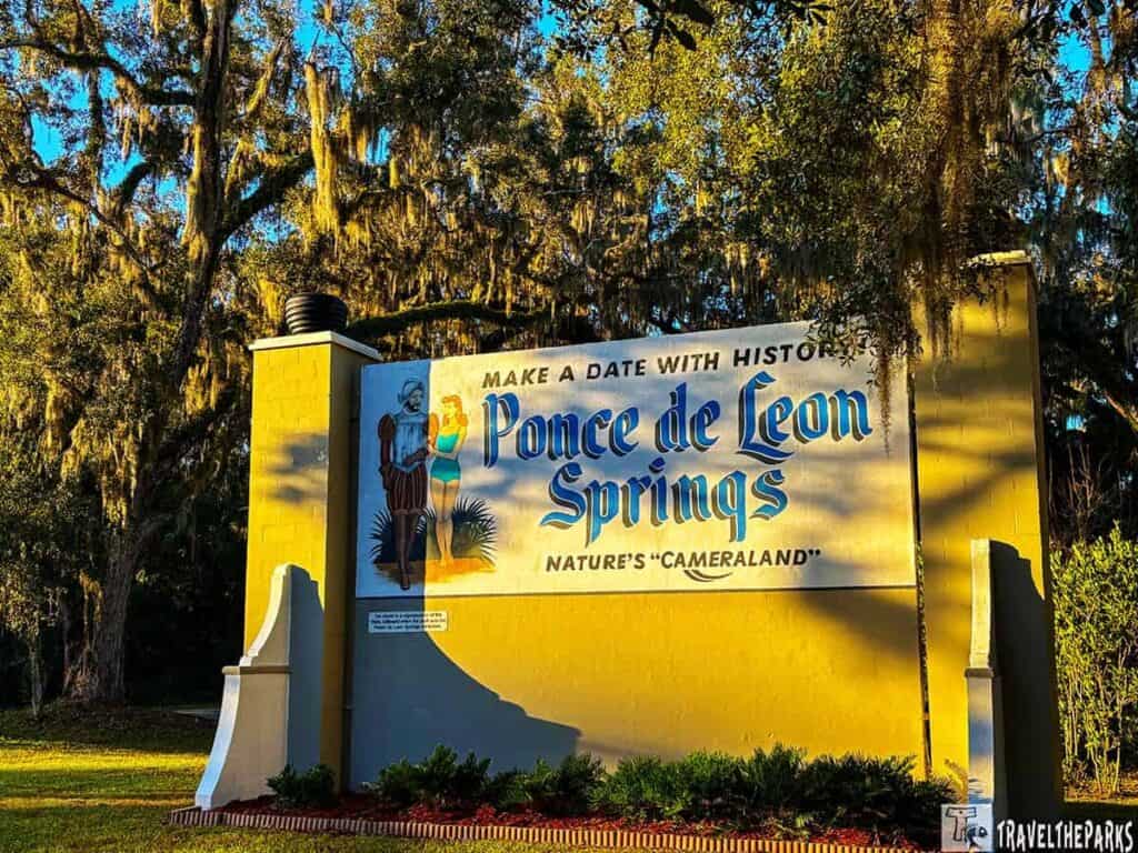 A sign for Ponce de Leon Springs with historical and swimmer illustrations, set against a backdrop of trees.
