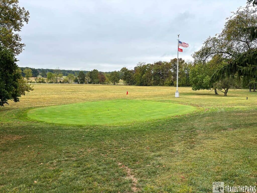 A circular putting green with a red flagstick on a grassy field at Eisenhower National Historic Site, surrounded by trees and a flagpole.